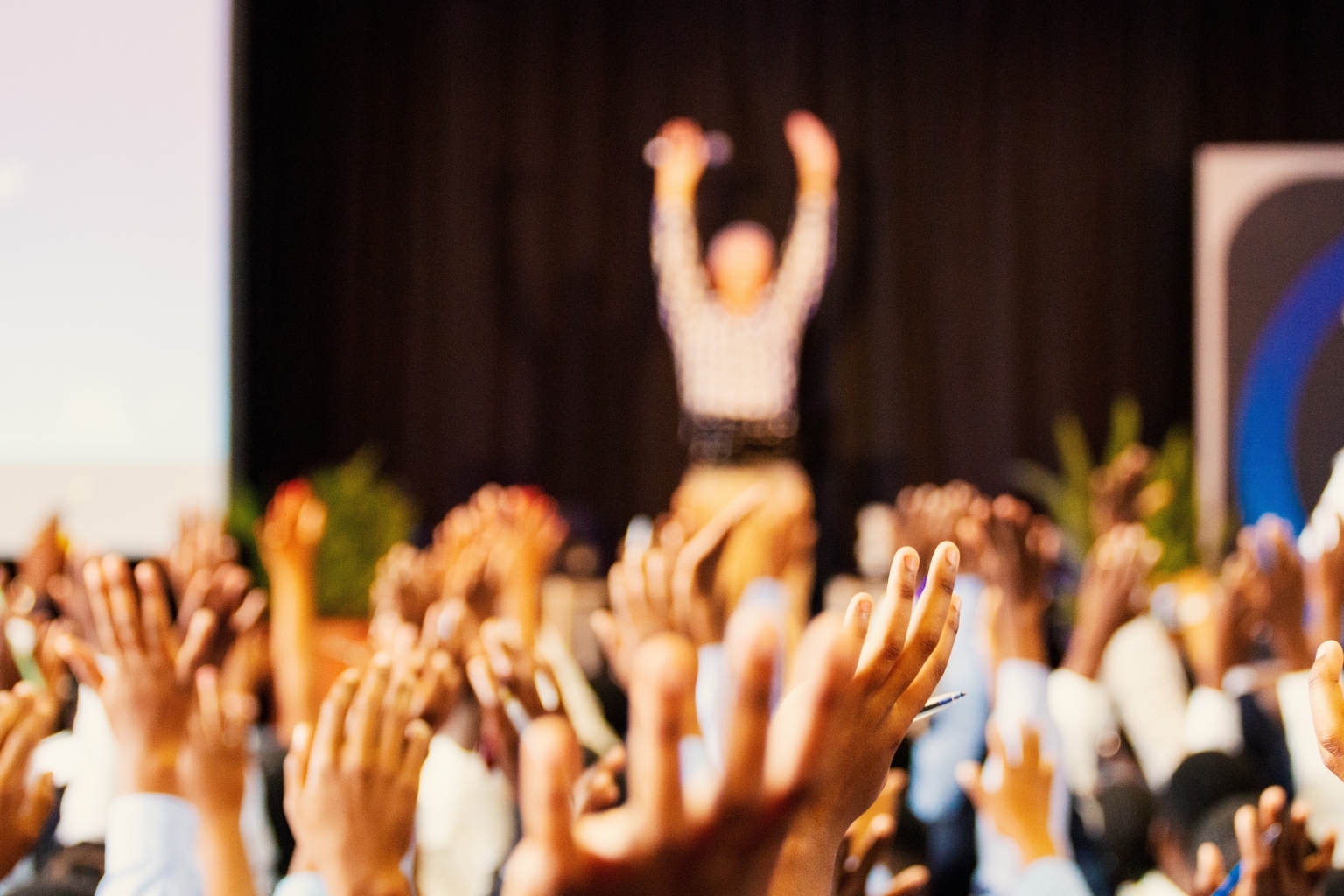 Hotel conference or banquet audience with hands raised toward a speaker on stage